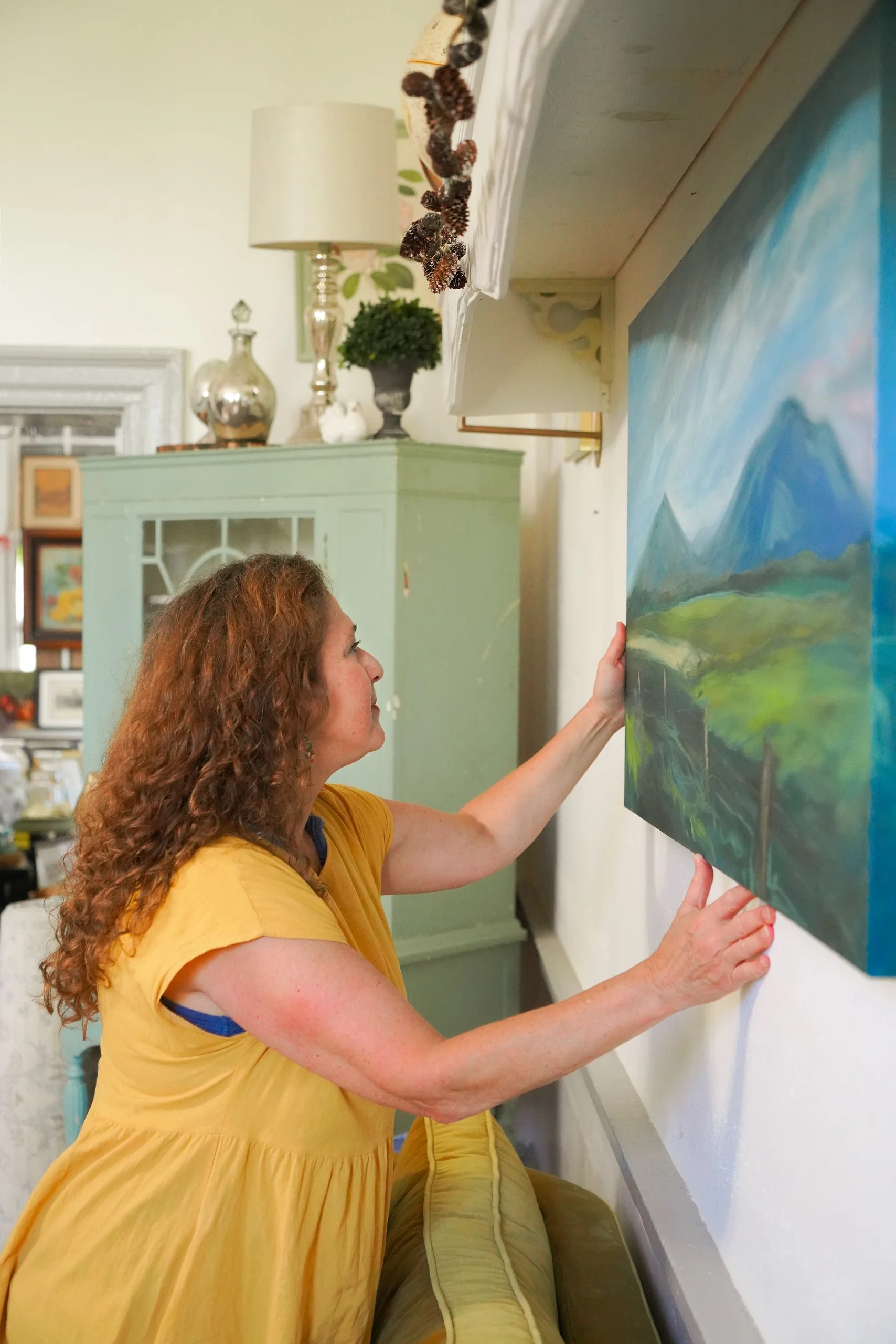 Woman in a yellow dress hanging a landscape painting.